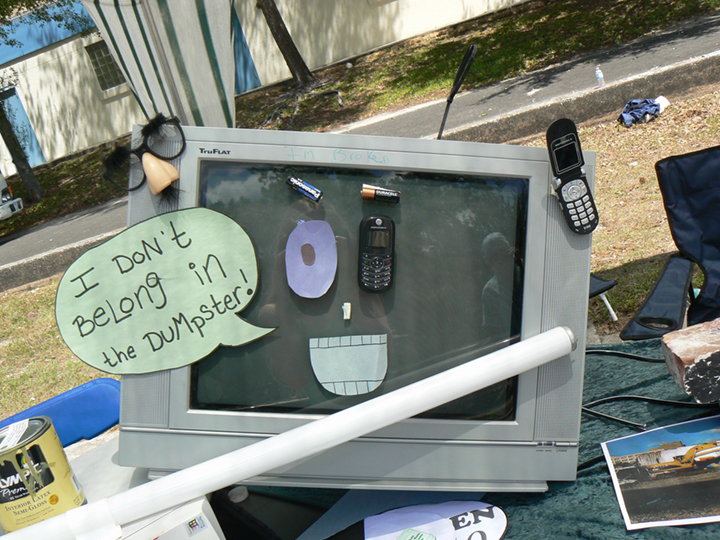 Art display: Old TV with cell phones and batteries forming a face on its screen, with a speech box saying 