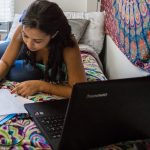 Student sitting on her dorm bed, looking at her laptop and doing homework
