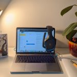 White study desk has a yellow lamp illuminating a laptop and headphone set. A small plant is set on the right corner of the table.