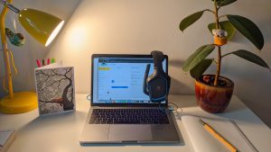 White study desk has a yellow lamp illuminating a laptop and headphone set. A small plant is set on the right corner of the table.